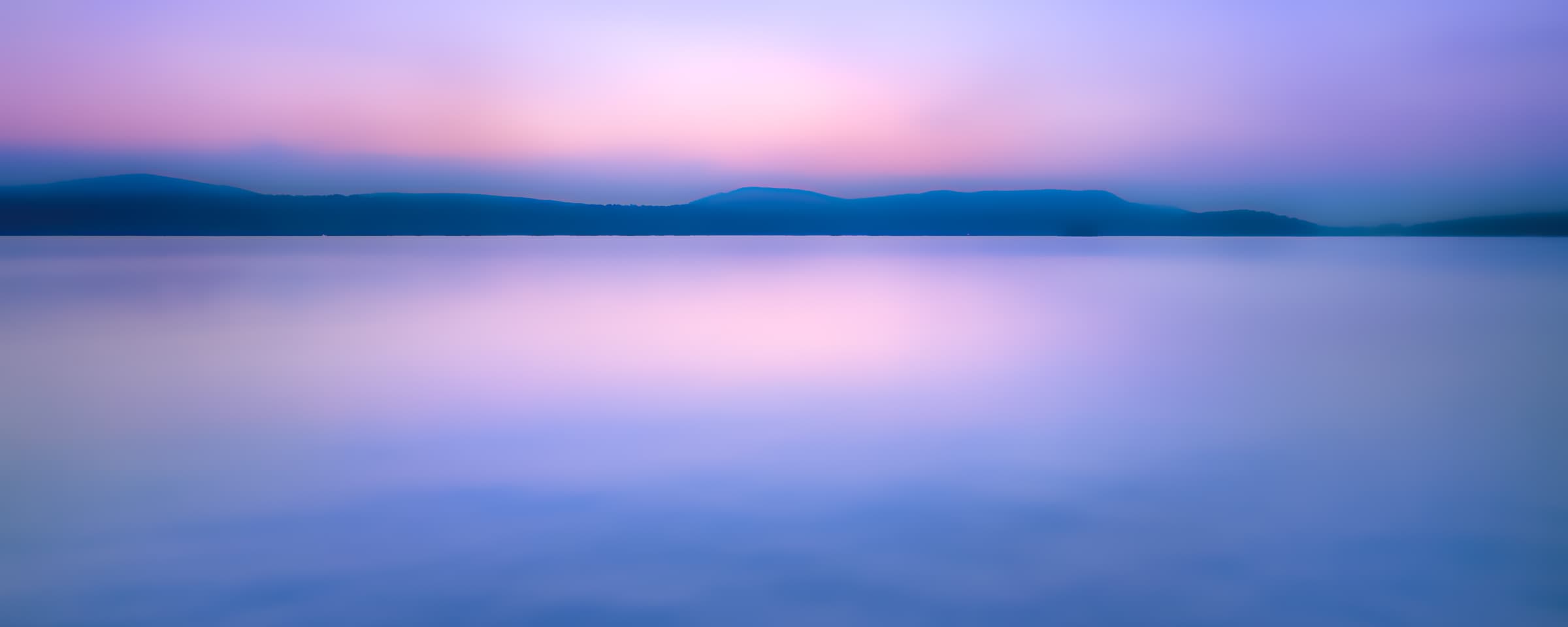 Fine art photograph of silky Adirondack lake waters beneath a pale pastel twilight sky. This contemplative image invites meditative reflection and a sense of timeless stillness.