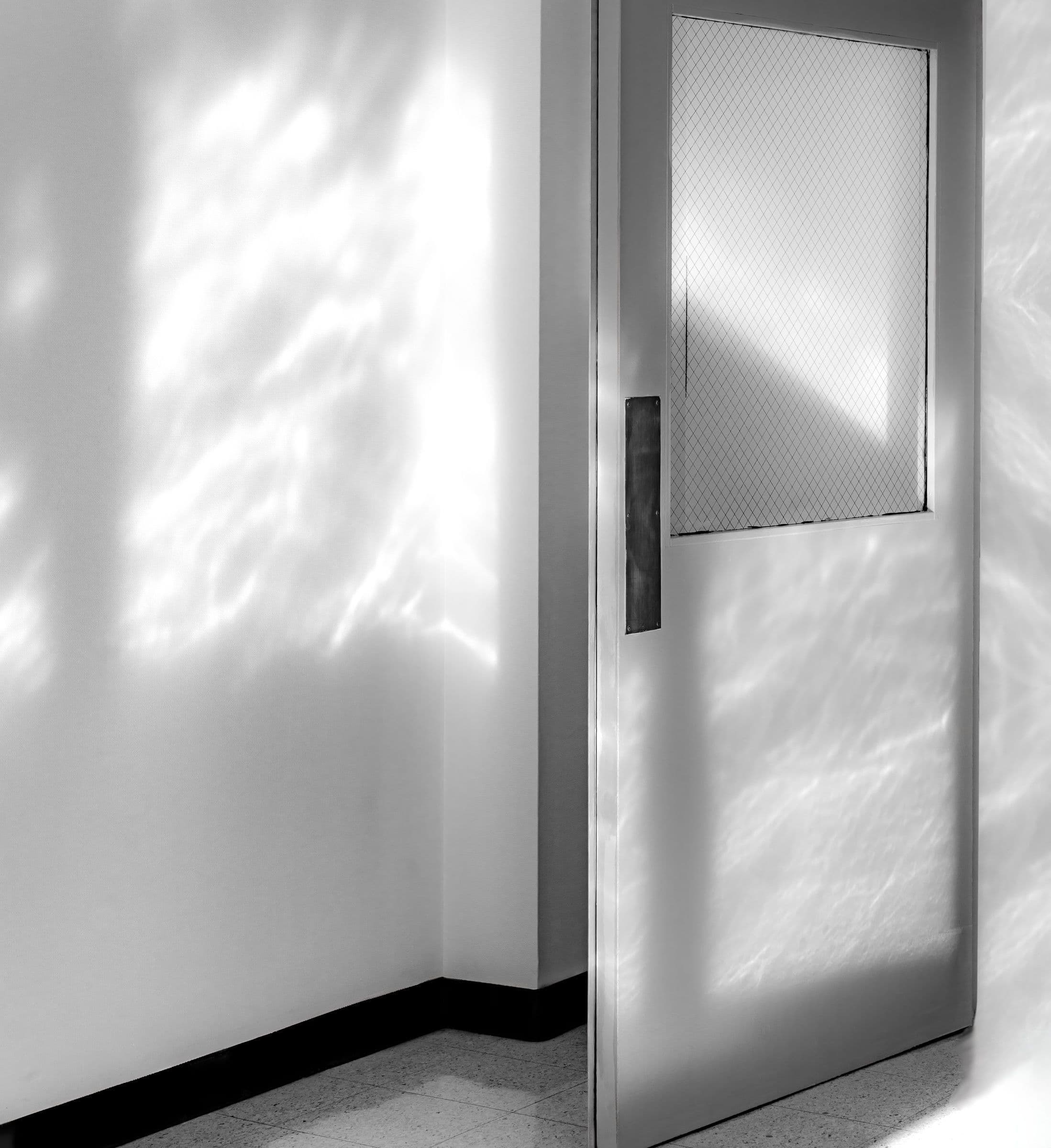 Black and white photo of an open door. Abstract light patterns shimmer on the wall and door in a quiet hallway.