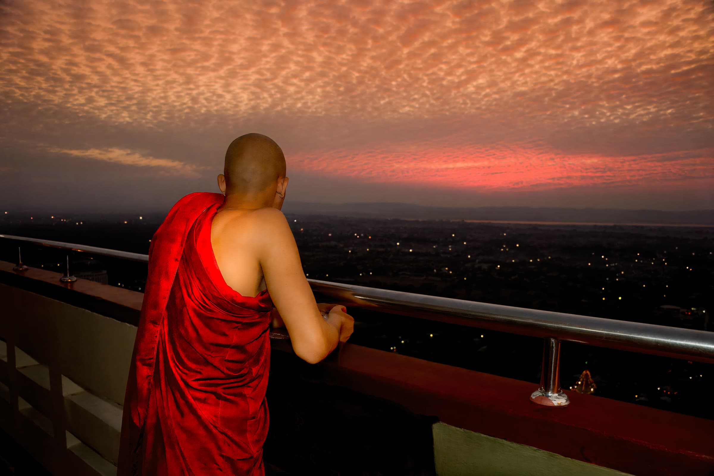 Sutaungpyei Pagoda, Mandalay Hill, Mandalay, Myanmar (Burma). Buddhist monk in red robe looks out from platform at darkening sky and cityscape below at twilght.