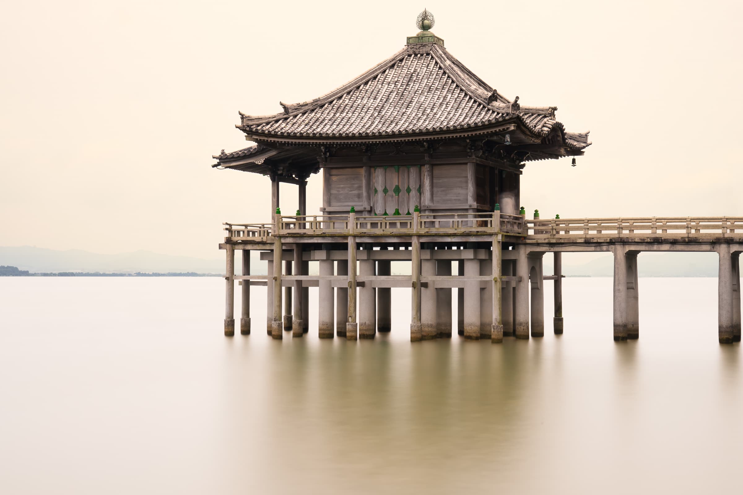 A Japanese pavilion appears to float above smooth, luminous water beneath a pale sky. The long exposure creates an atmosphere of stillness and contemplation.