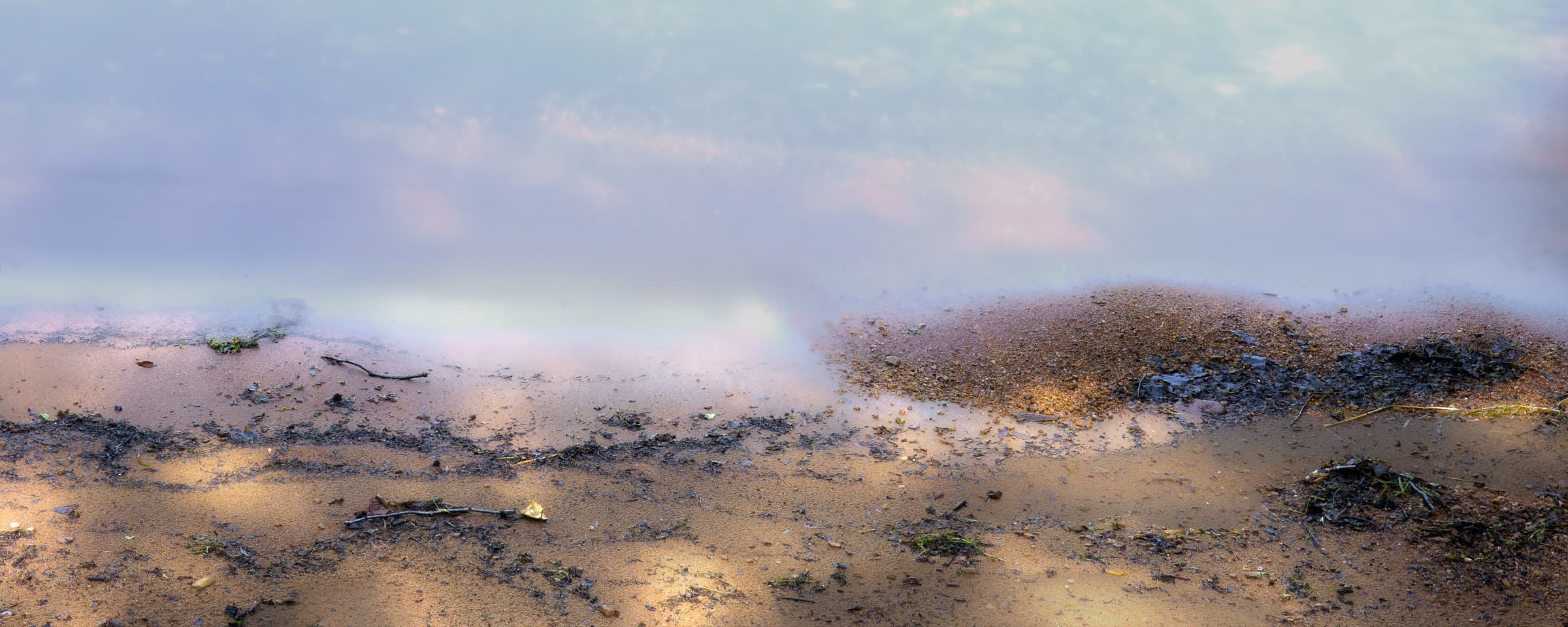 Soft blue and pink cloud reflections color shallow water beside a sandy shoreline. Shifting light and a blurred waterline soften the boundary between land and lake.