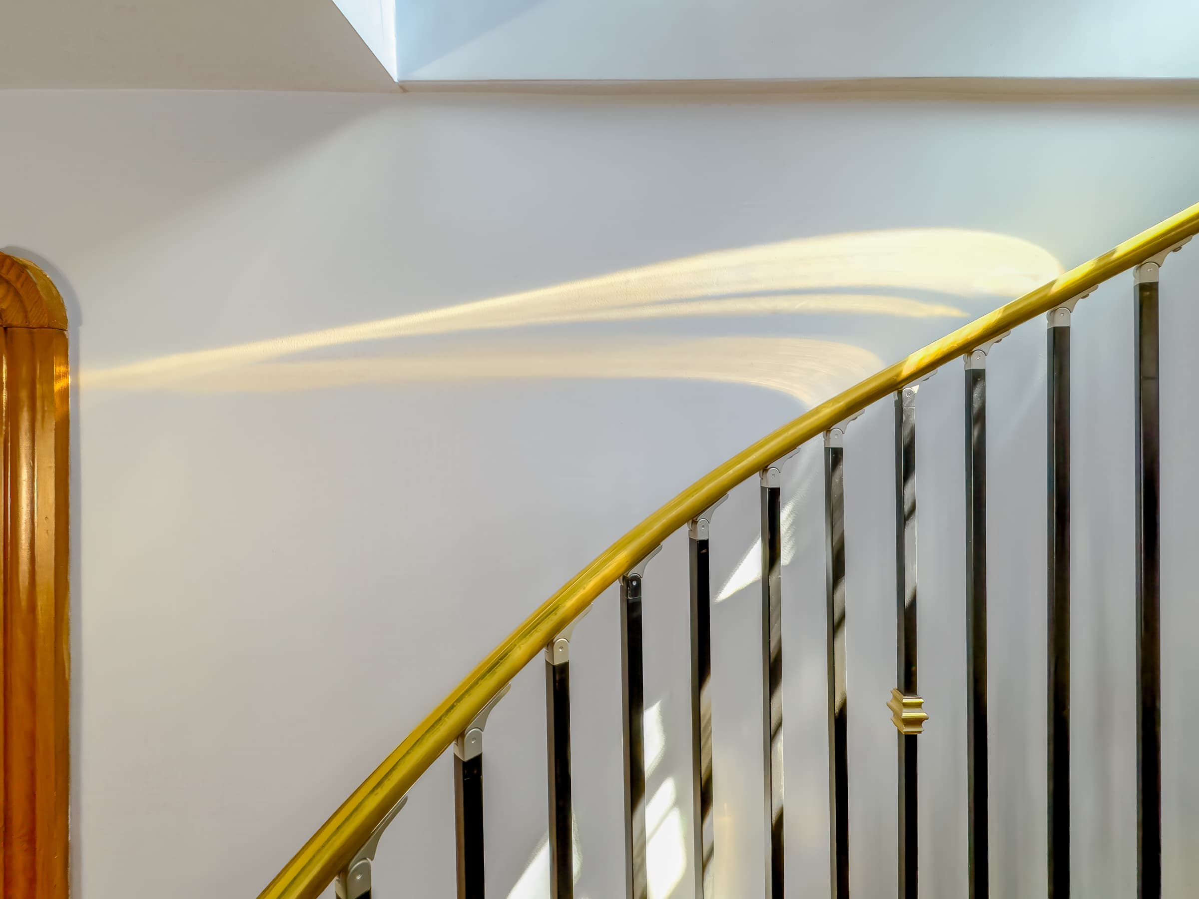 A quiet hallway photograph of a curved staircase banister with abstract light arc on the wall, creating a minimalist composition. The curve of light interacts and contrasts with dark vertical railings.