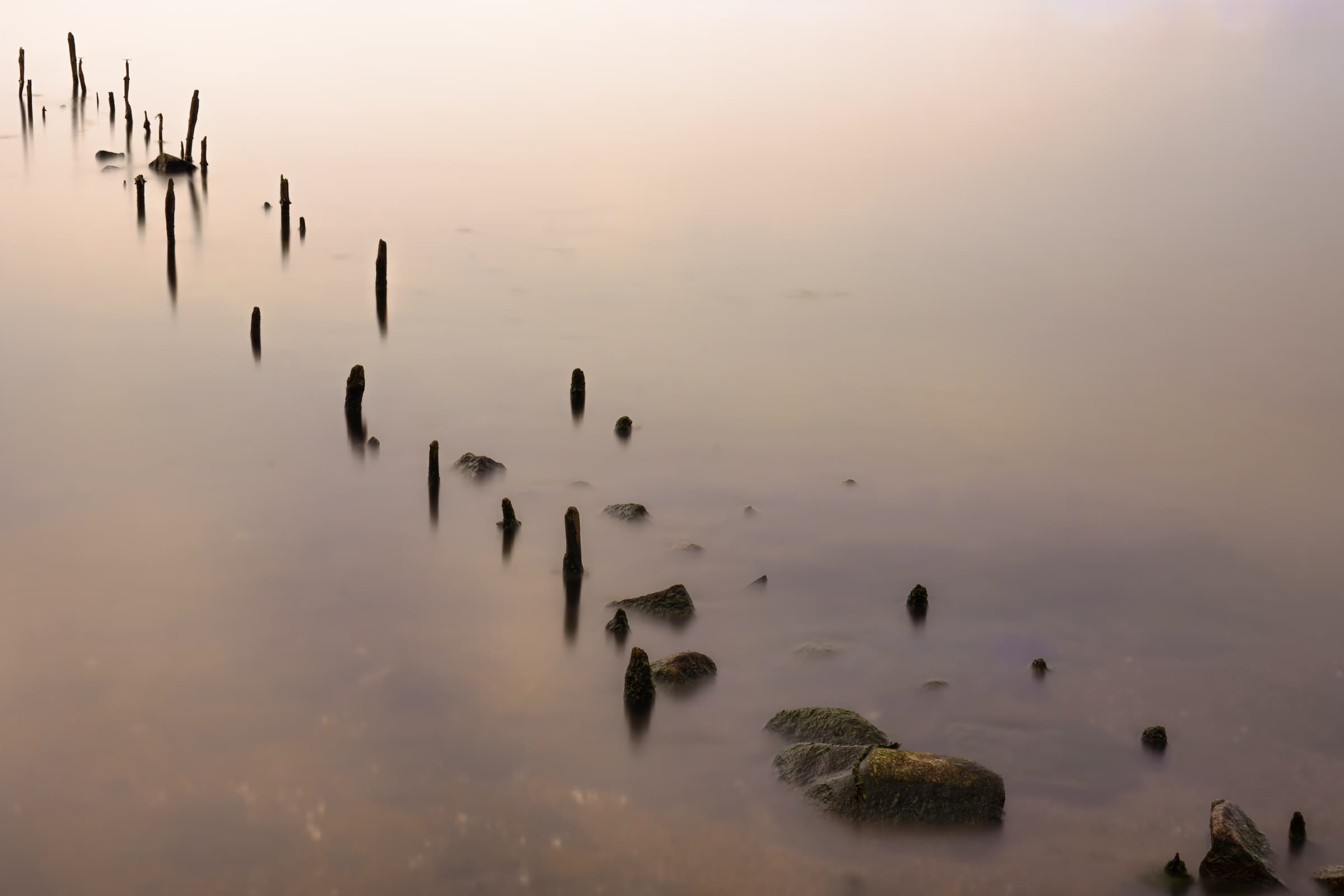 Weathered wooden posts and scattered rocks stretch diagonally through softly colored water in a long-exposure photograph at Lake Biwa, Japan.