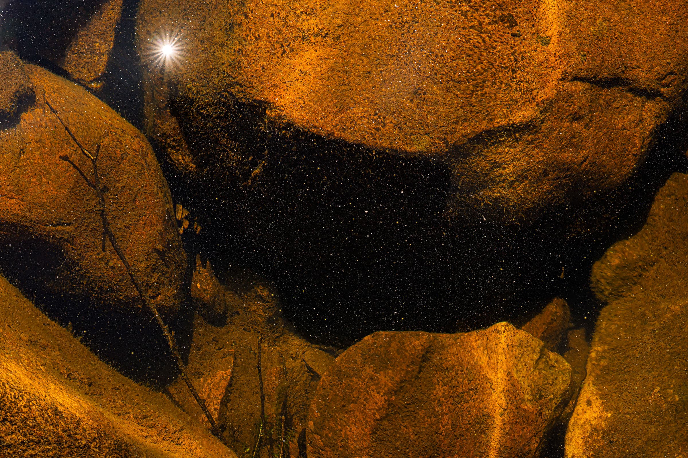 Photograph of golden rocks beneath water with the surface showing sunlit reflections and specks like stars. The image evokes a contrasting abstraction of cosmic and terrestrial. 