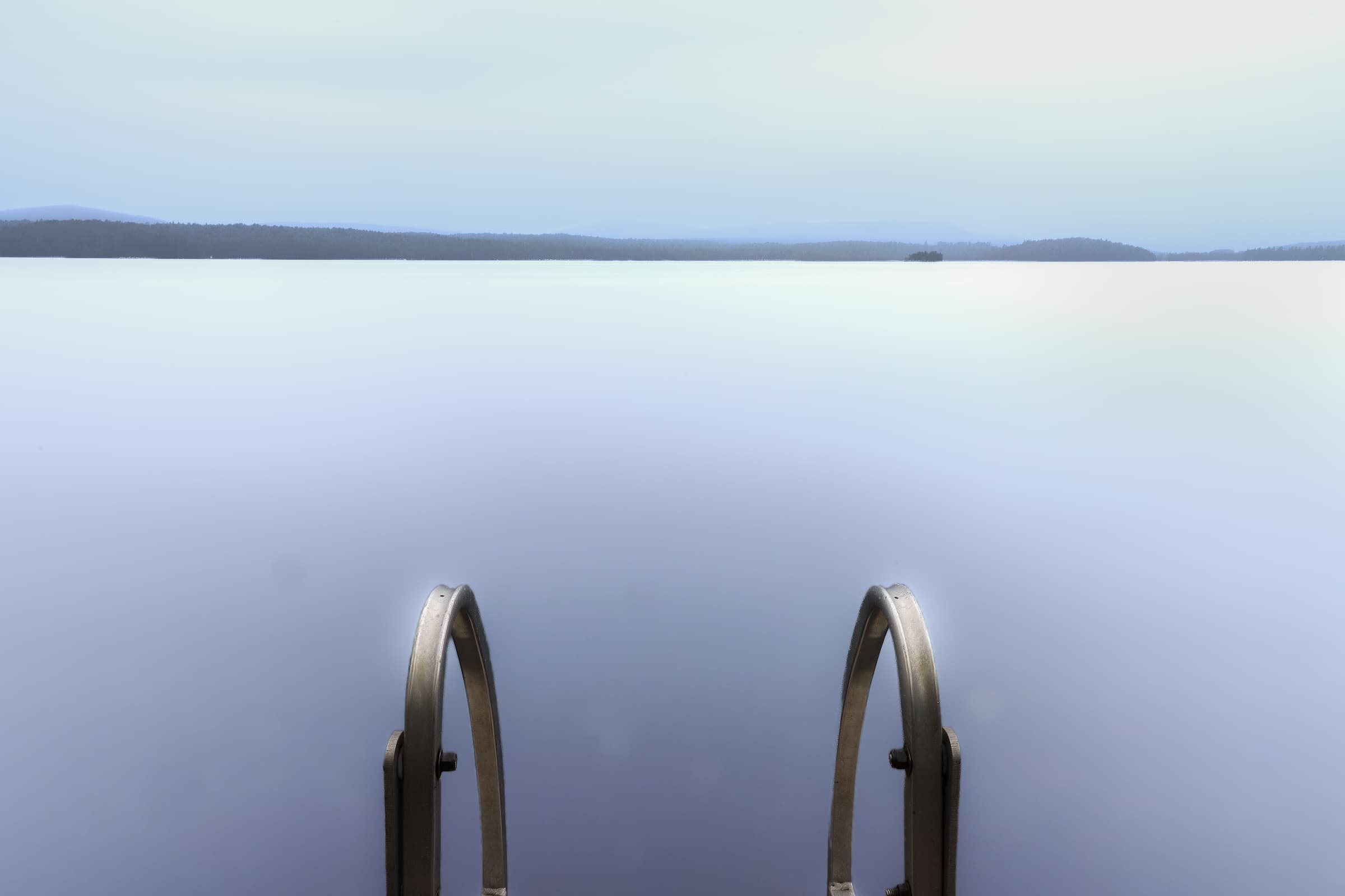 A metal swimming ladder descends into a smooth pale-blue lake, with distant low mountains beneath a hazy sky in the distance.