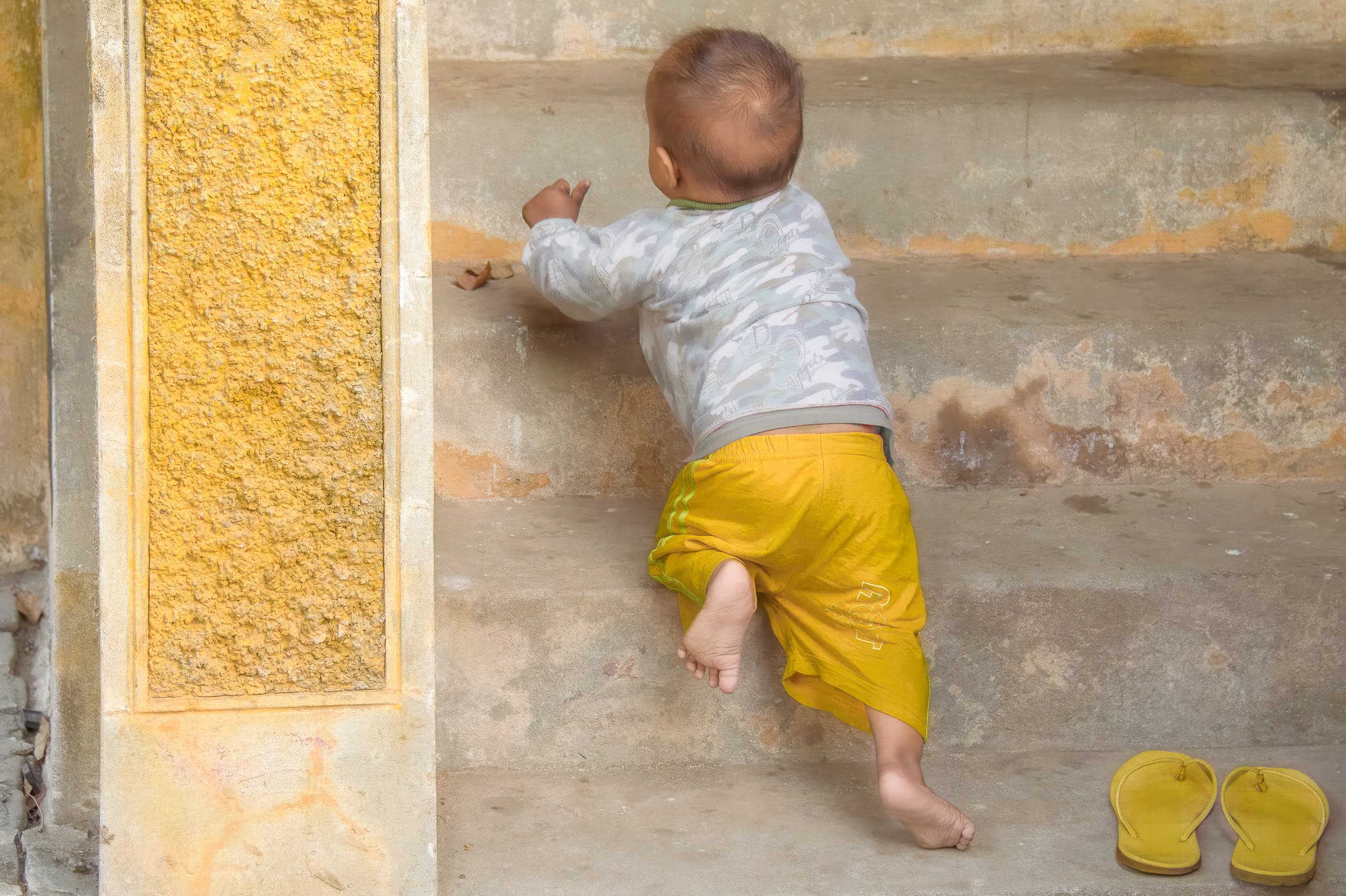 Confident barefoot toddler climbs old stone steps. Warm hues and bright yellow dominate.