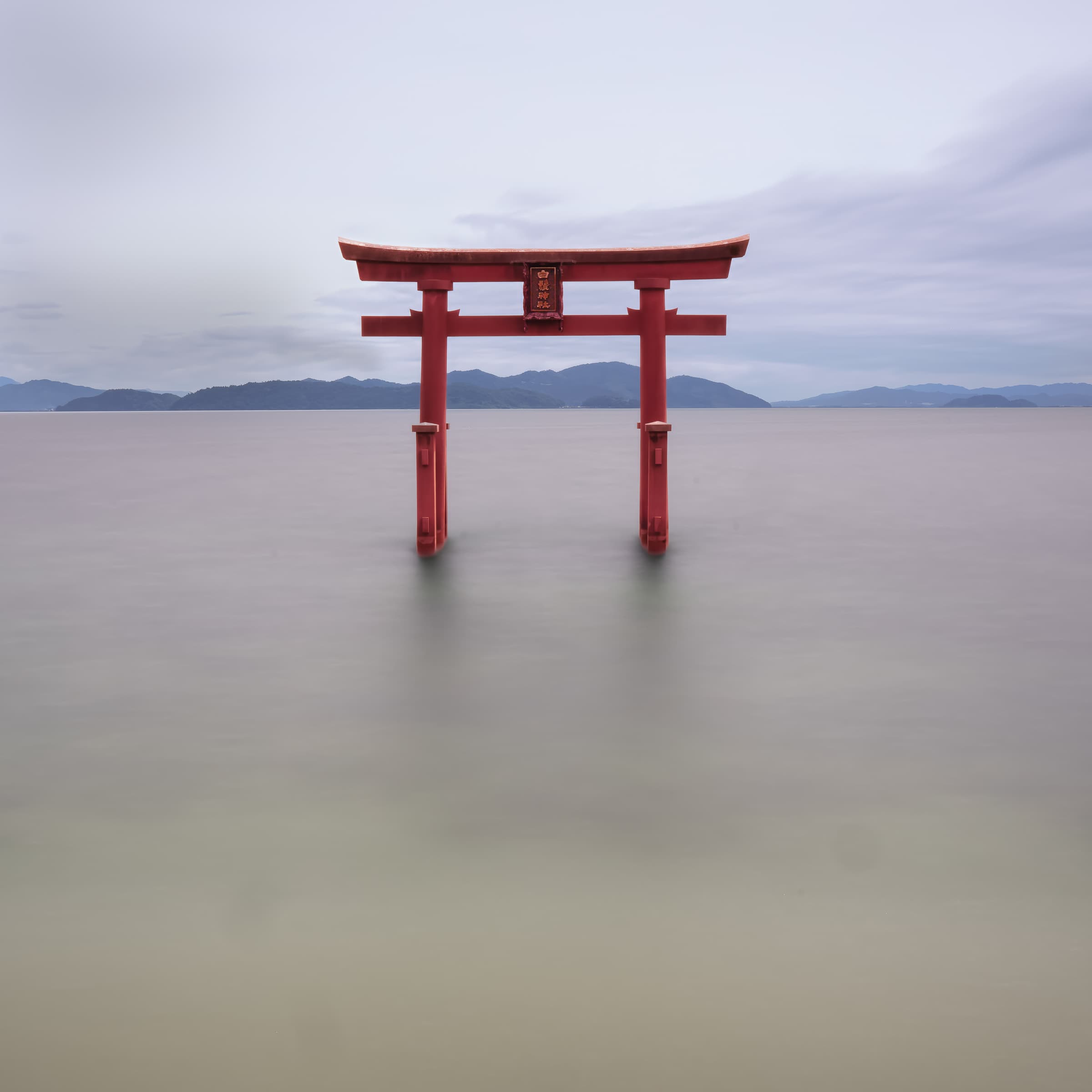A red torii gate stands in smooth water beneath a pale sky. Distant blue mountains are visible in the background.