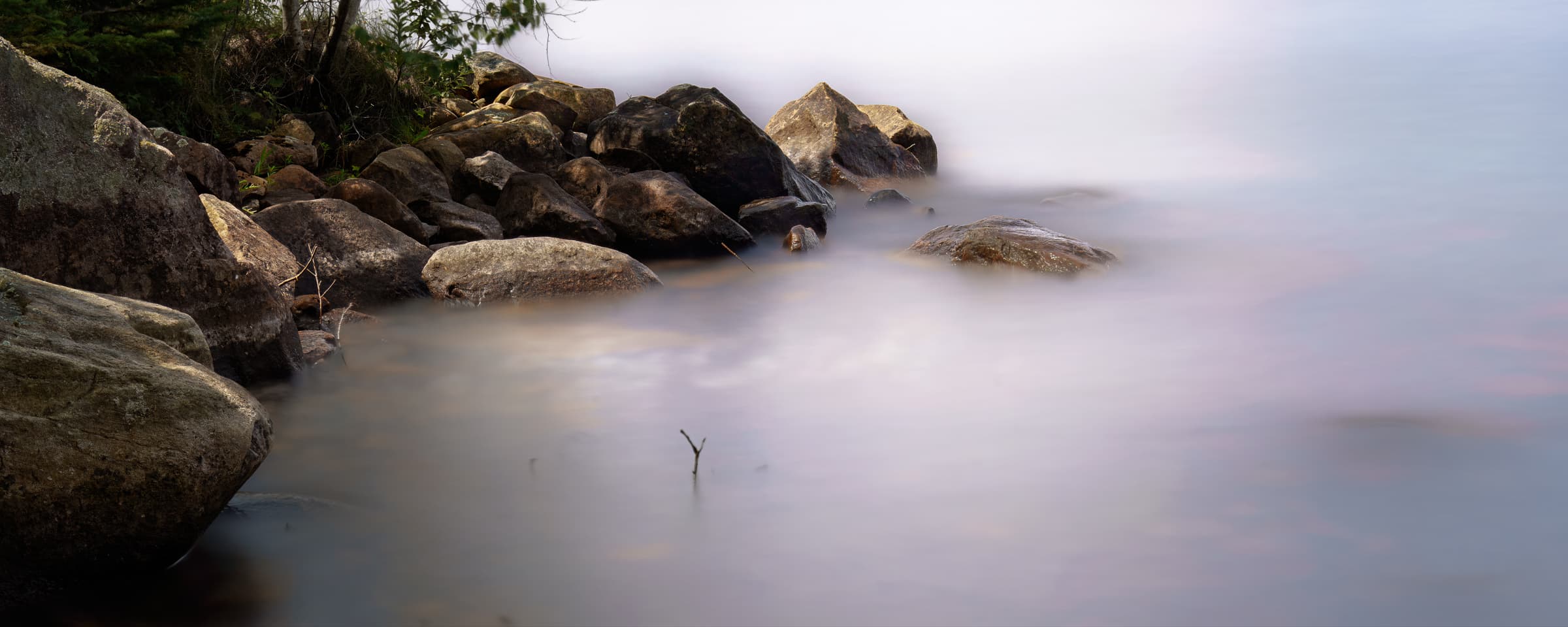 Large shoreline rocks sit at the edge of a lake while long-exposure water blurs into a soft mist around them. The image emphasizes the contrast between solid stone and flowing water.