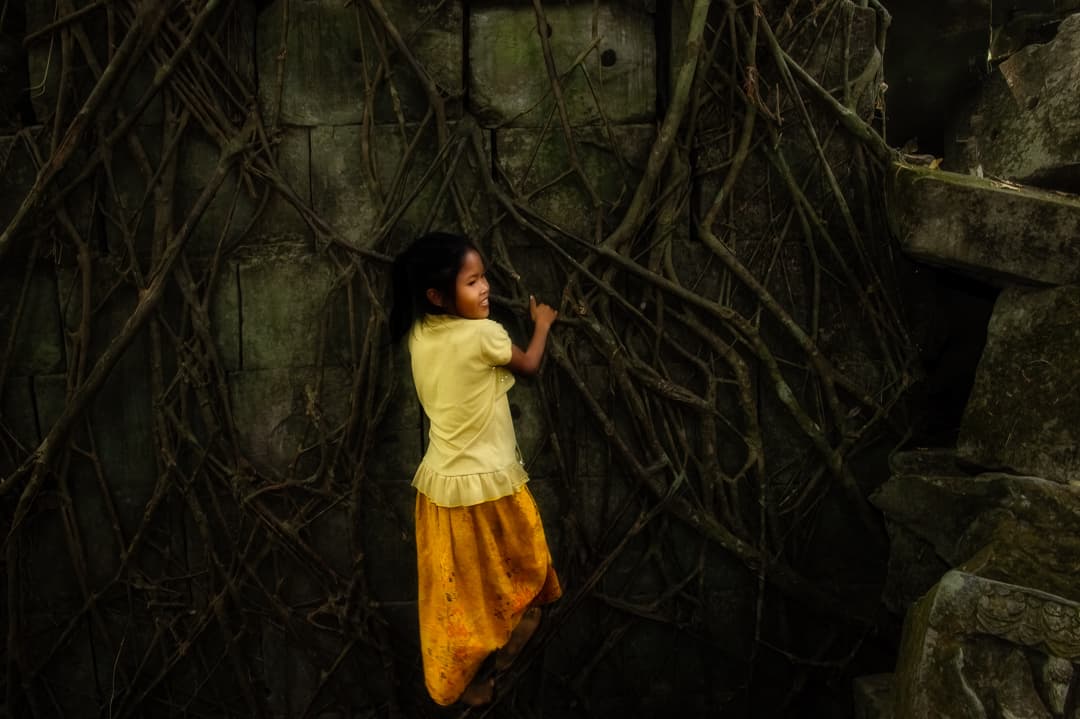 Beng Mealea Temple, Angkor Archeological Park, Siem Reap, Cambodia. Daring young girl in orange and yellow climbs tangled roots on dark rock wall of temple ruins.
