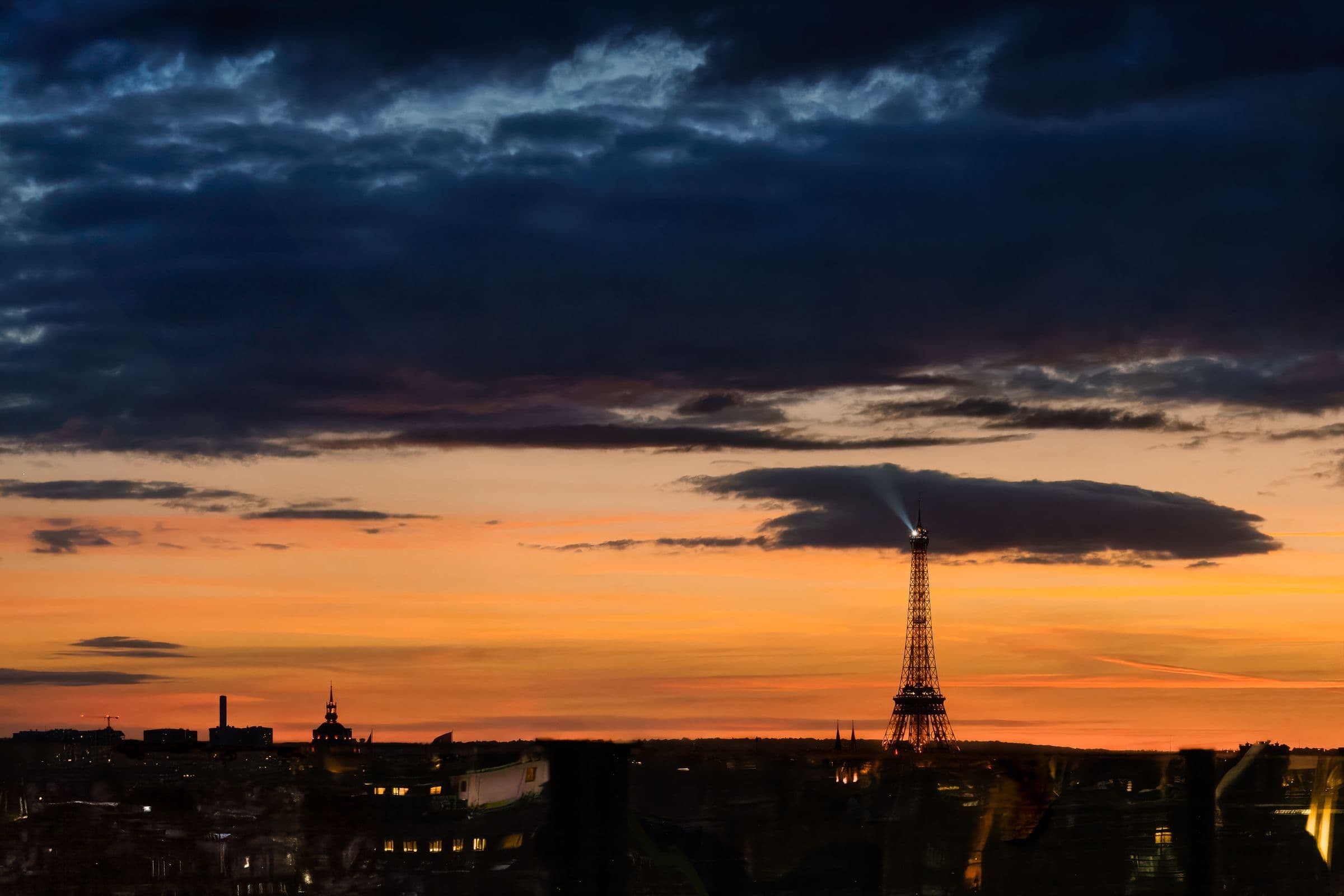 Fine art photo of the Eiffel Tower at sunset, with a dramatic sky, city reflections, and a glowing horizon capturing the moody romance of twilight in Paris.