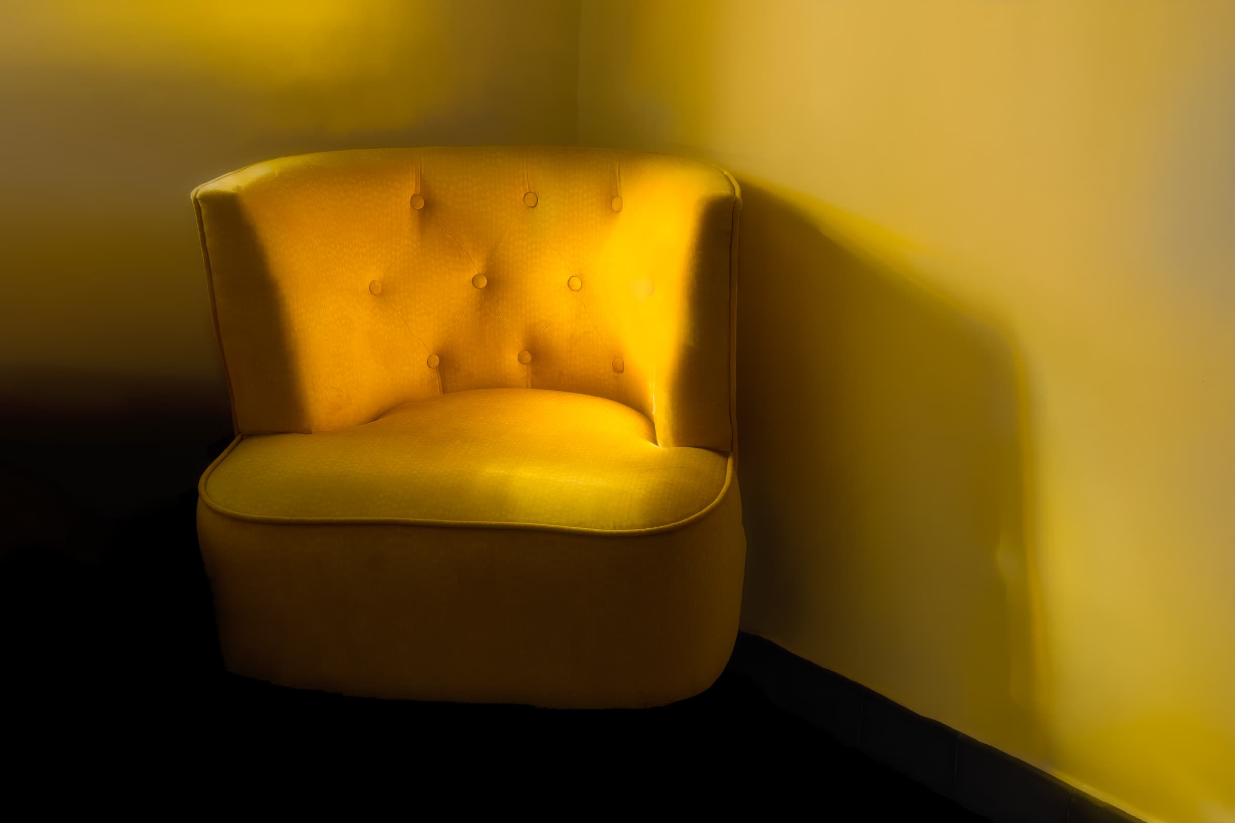A yellow chair is illuminated by golden sunlight as it rests in the corner of a room. Soft light and gentle shadows spill across the chair and wall