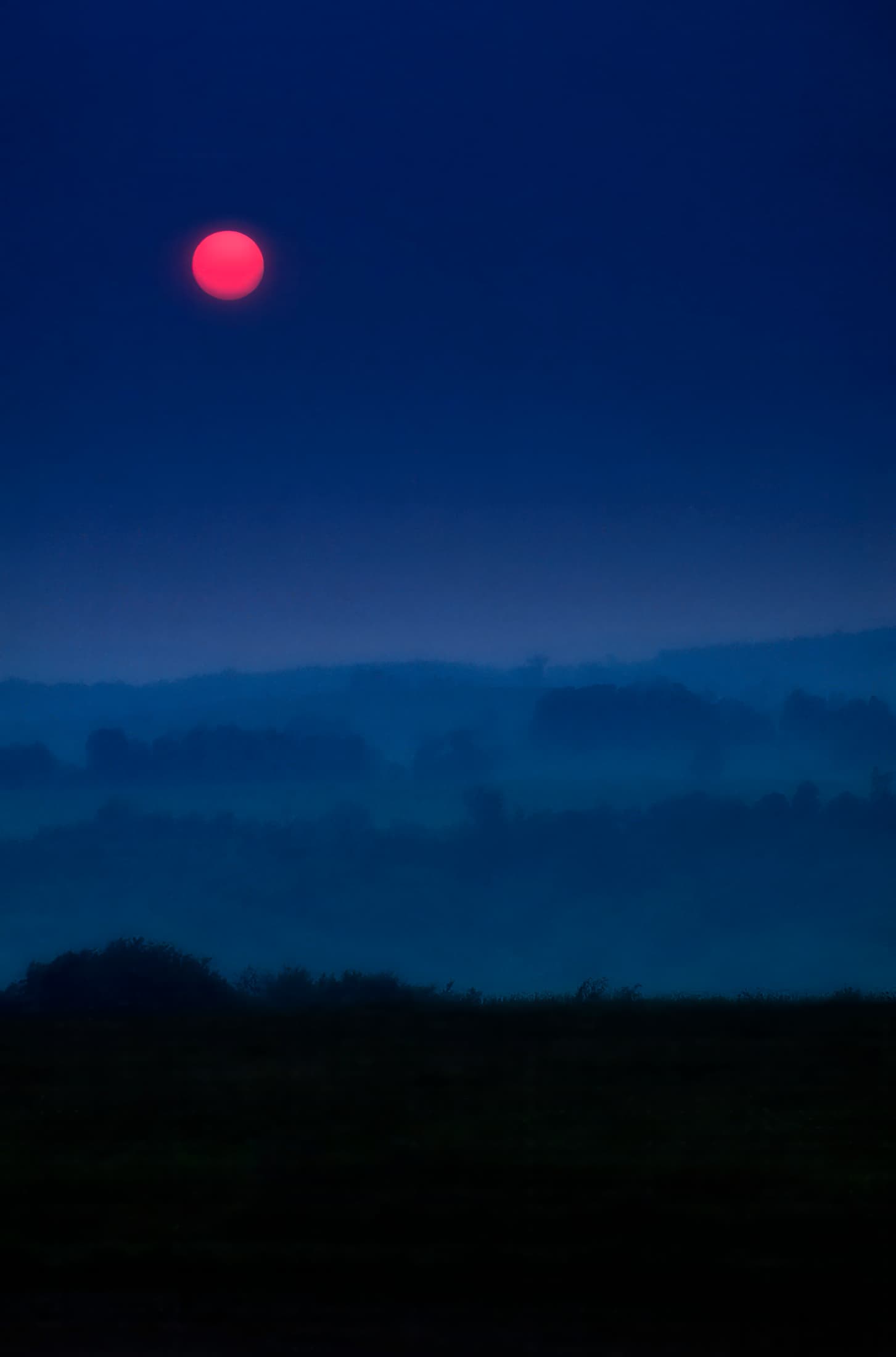Layered landscape with dark foreground, blue and hazy distant hills, dark purple-blue sky with red sun. 