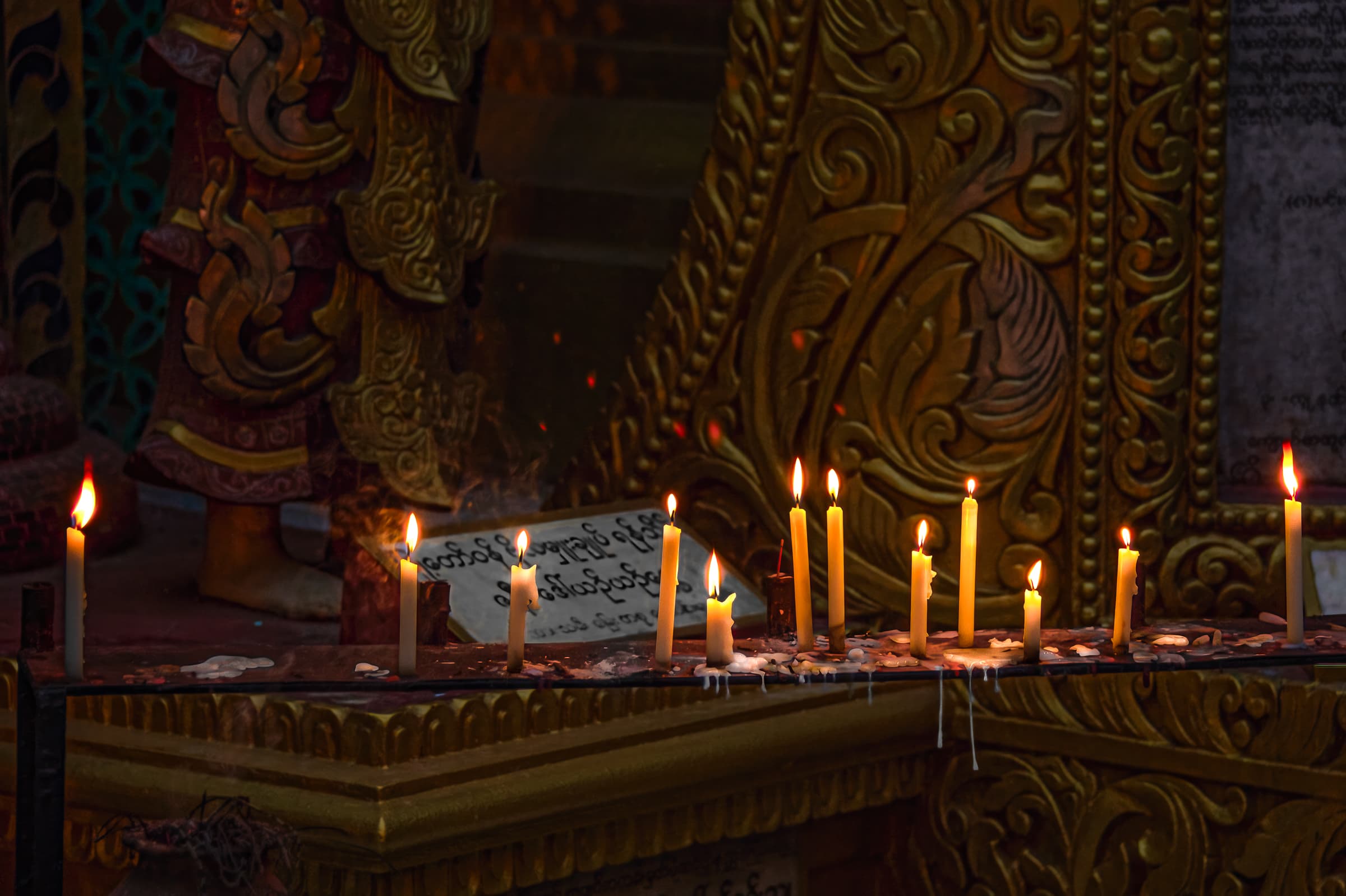 Mandalay, Myanmar. Twelve thin candles burn on slab in front of intricately carved details on wall in Buddhist temple of Sutaungpyei Pagoda on Mandalay Hill.