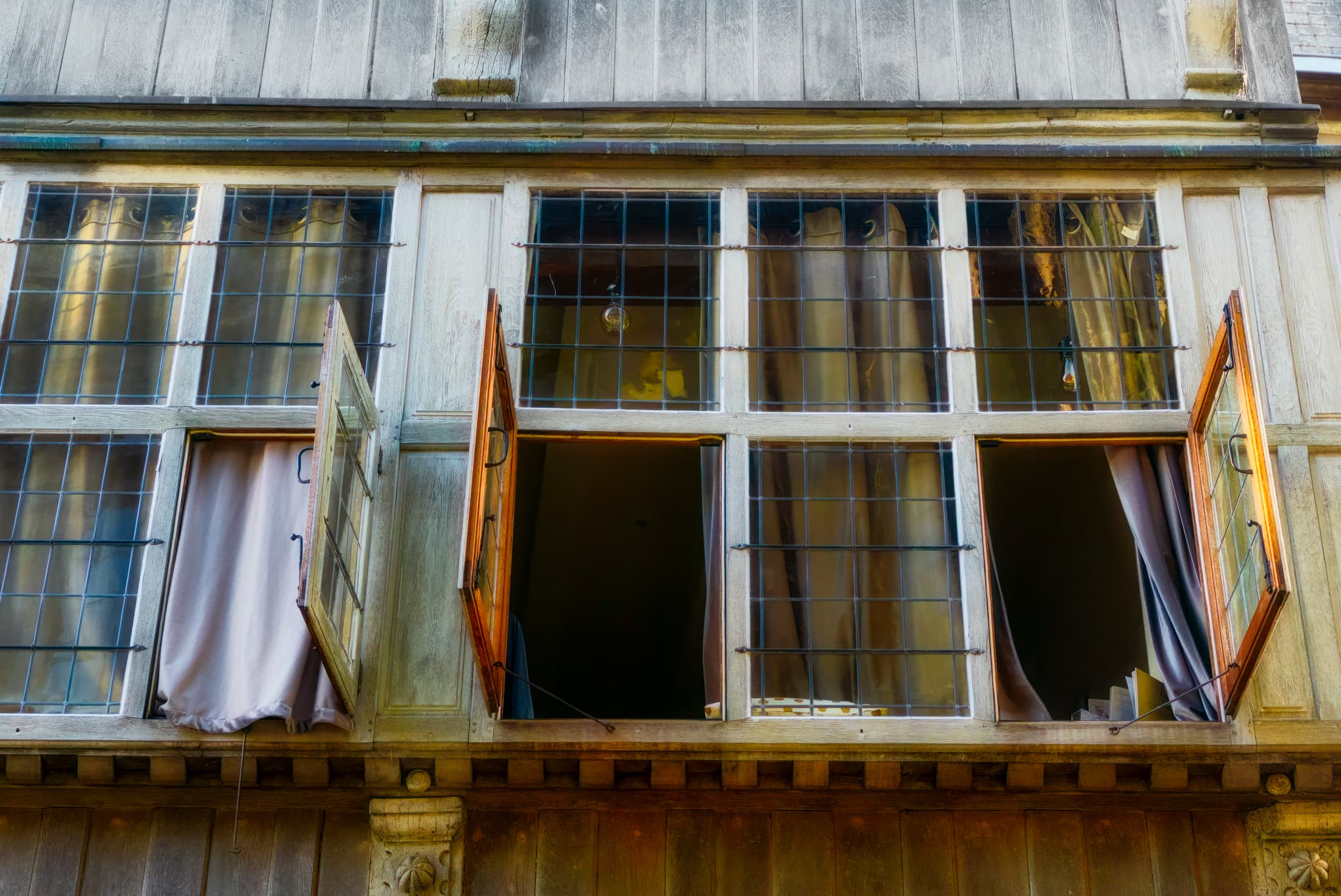 Open windows in an old building reveal soft curtains spilling outward, framed by worn wood and gridded panes. The photograph evokes timeless mystery and quiet elegance.