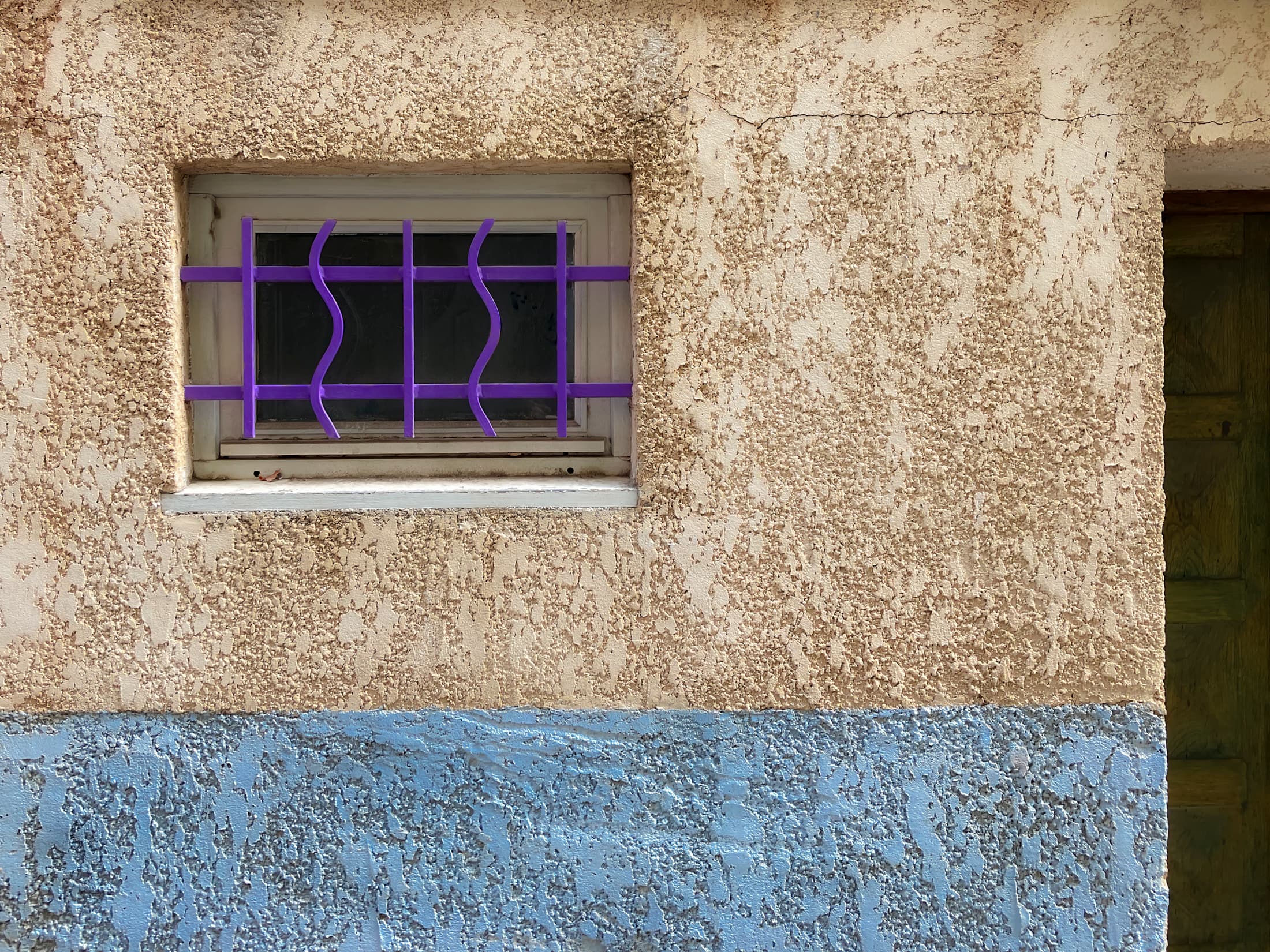 Collioure, France. Window and part of a door set into textured building wall, creating a multi-colored geometric composition. The purple bars on the window and closed door suggest a private place with a sense of contentment.