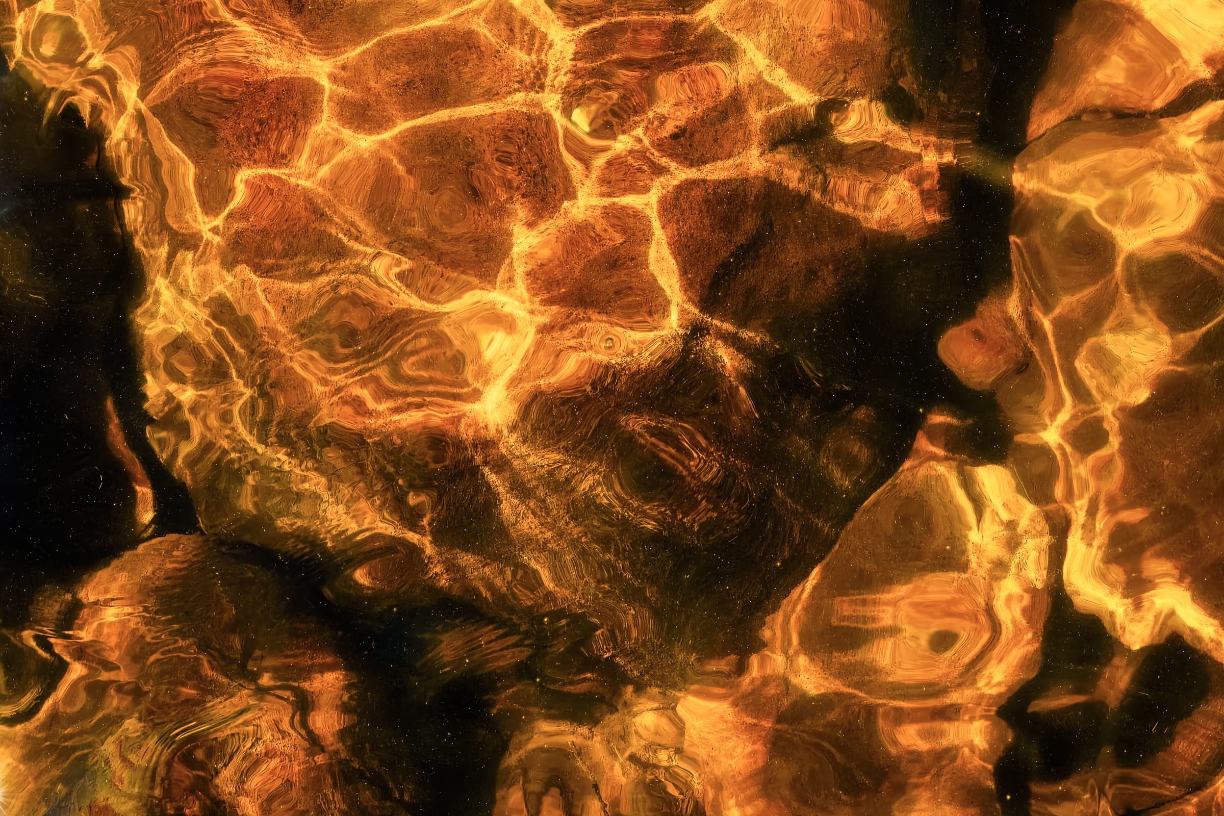 Underwater view of rocks lit by intense golden sunlight, with rippling patterns of light and shadow across their surfaces. Abstract photograph emphasizing transient beauty and joyful, momentary flashes of gold.