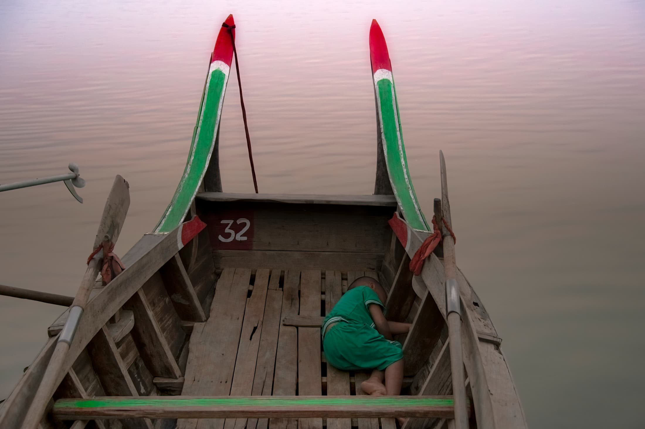 Taungthaman Lake, Mandalay, Myanmar (Burma). Young child sleeps in wooden boat with green and red markings on quiet water, its prow pointed towards soft rose-colored light.