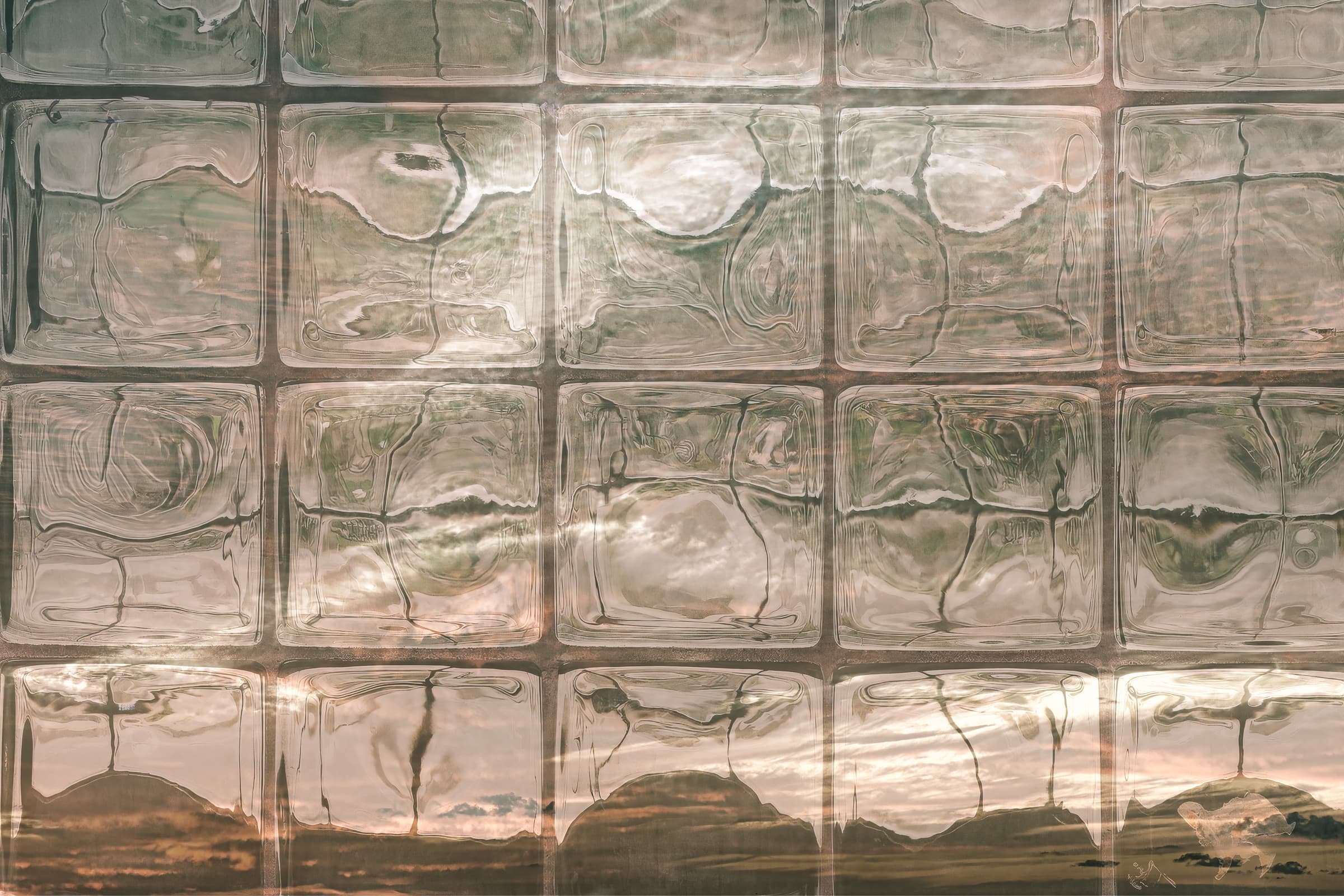 Grid of glass blocks filled with distorted reflections and warm sunrise tones, suggest hills and clouds inside the squares. Abstract fine art photograph emphasizing light, texture, and organic forms within a rigid structure.