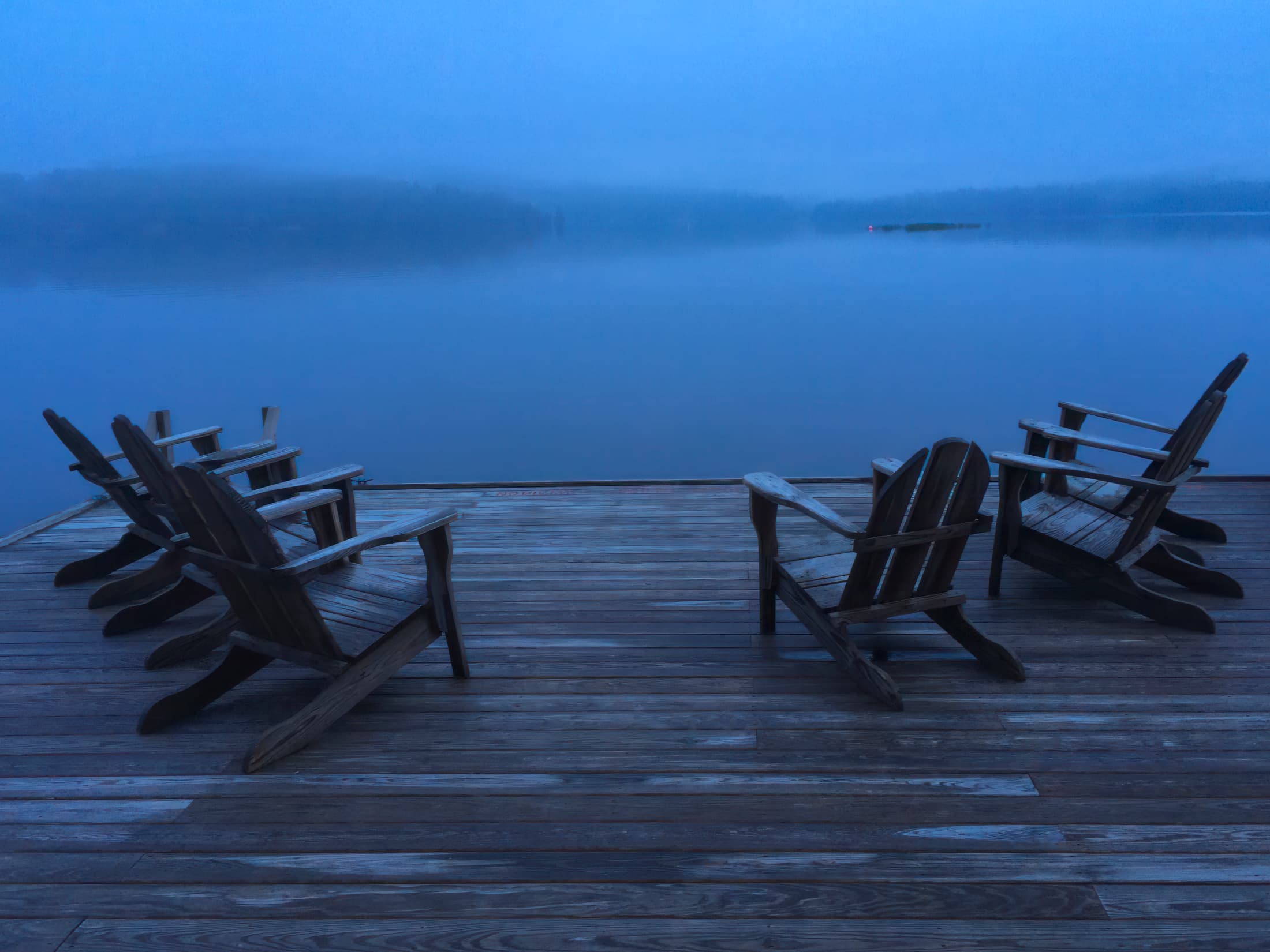 A group of empty Adirondack chairs sits on a wooden dock facing a calm, misty lake under the blue glow of twilight. The scene is tranquil and contemplative, inviting quiet reflection.