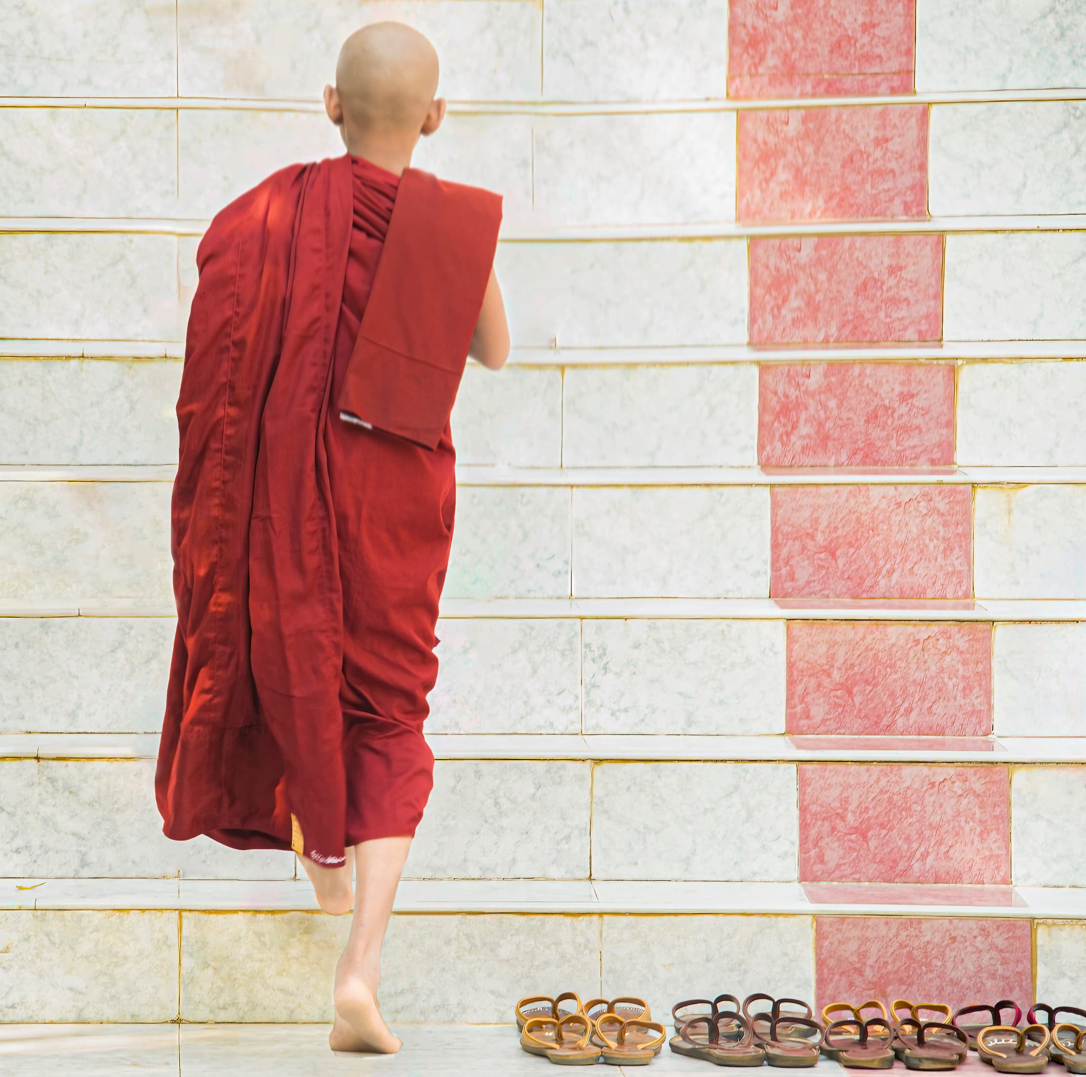 Mya Set Kyar Nunnery, Mandalay, Myanmar. Boy, young monk in red robe walks up white marble steps to temple.