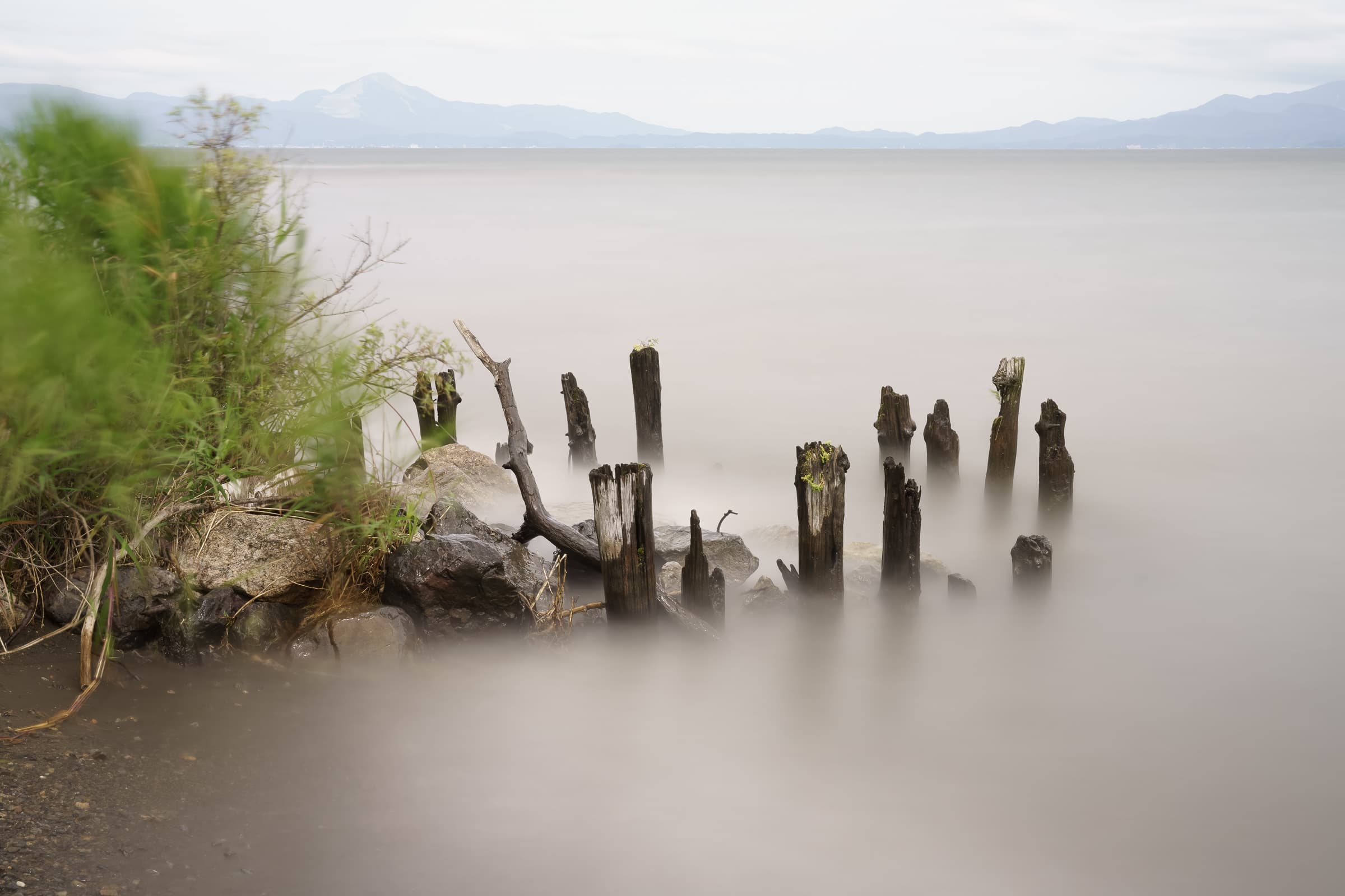 Weathered wooden pilings rise from silky long-exposure water along the shore of Lake Biwa, with hazy mountains in the distance.