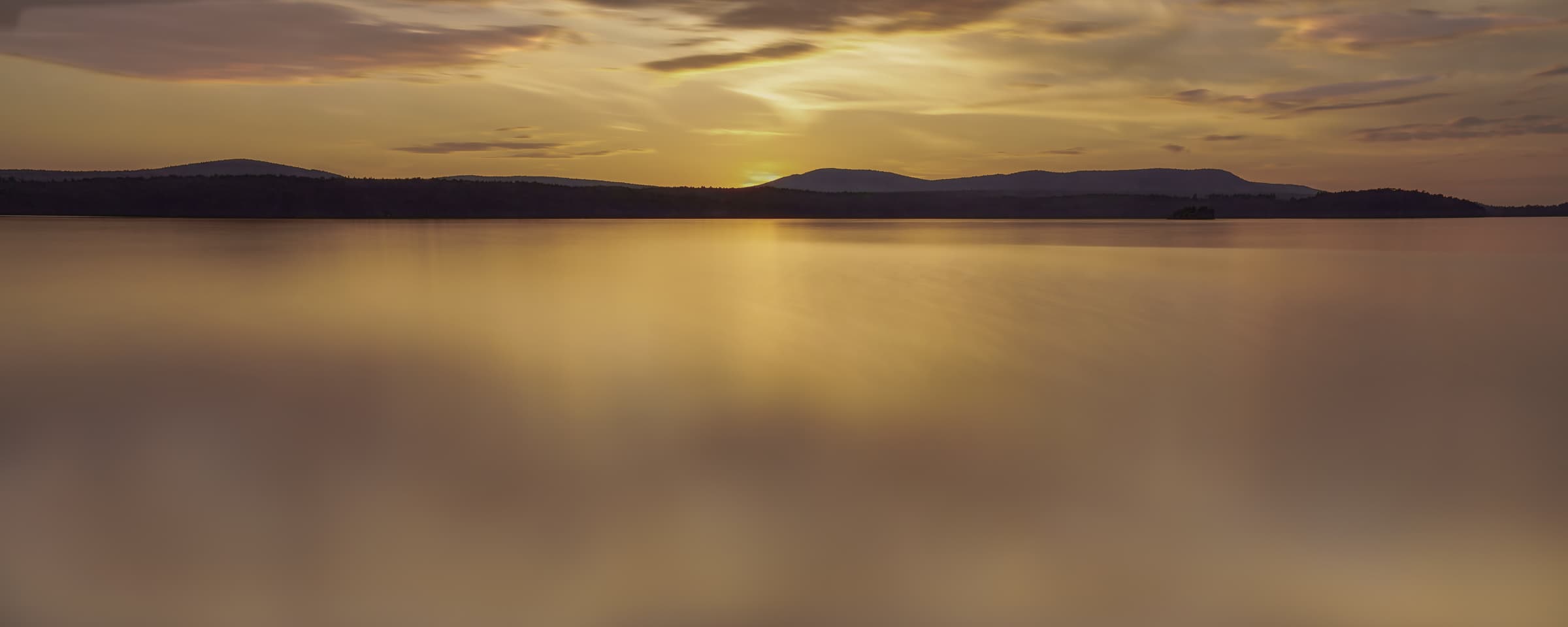 Golden sunset light spreads across a lake beneath a shadowed line of distant hills. Long exposure softens the water into shimmering areas of light and shadow.
