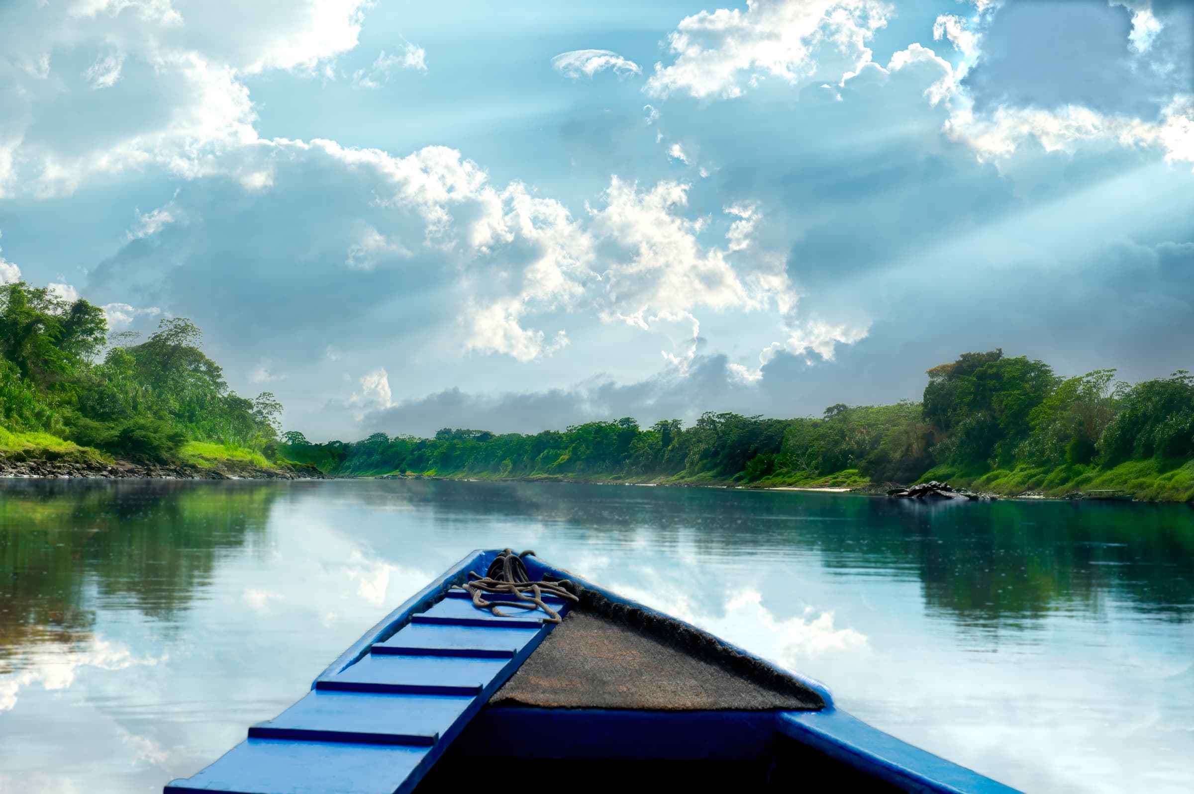 Tambopata National Reserve, Peru. Clouds with light beams reflect in water as boat travels up the Tambopata River. Rainforest trees and plants line the riverbanks.