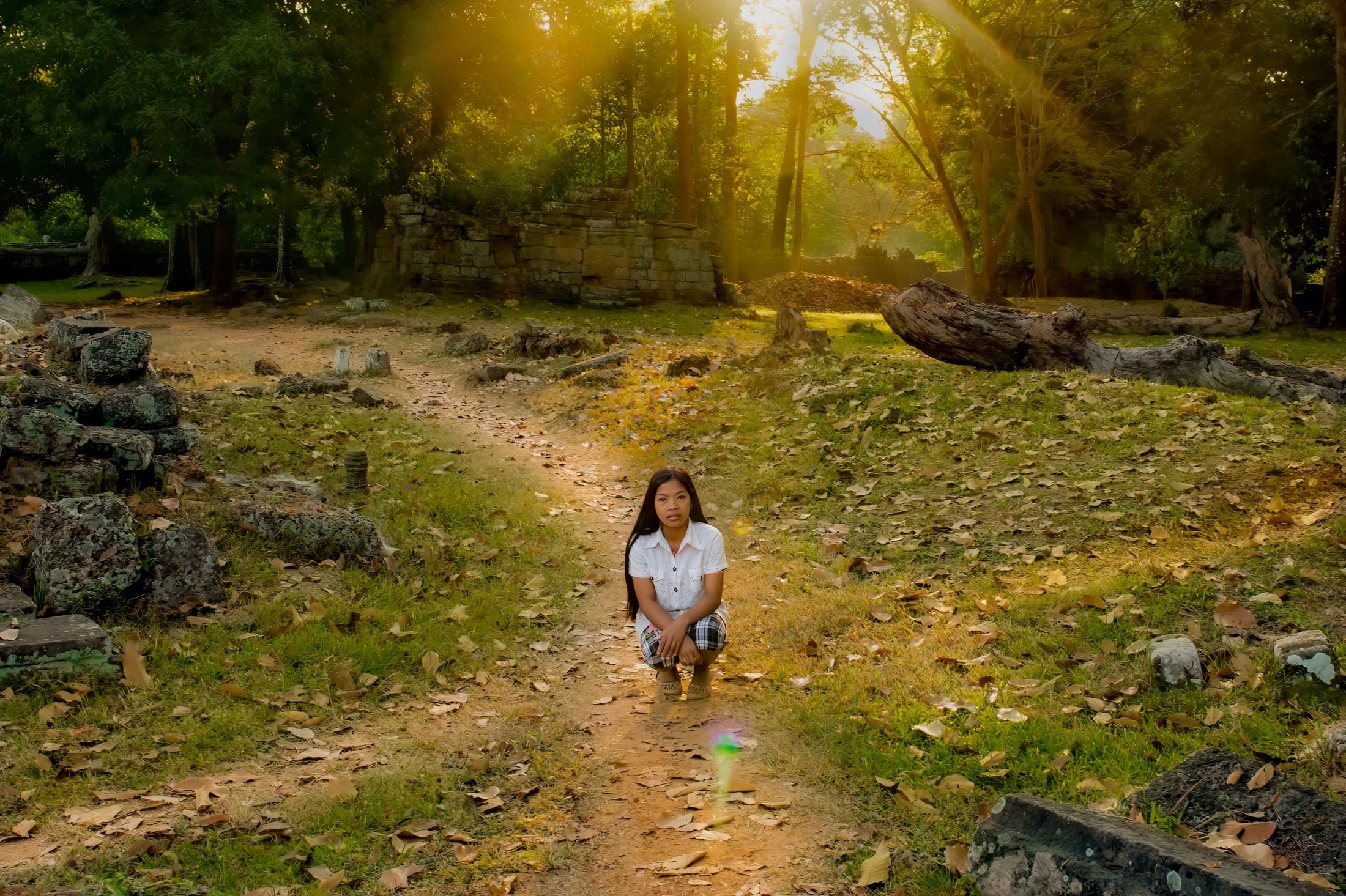 Banteay Kdei Temple, Siem Reap, Angkor Archeological Park, Cambodia. Sun rays stream in background as teen girl sits at crossroads of paths on temple grounds. Strewn stone blocks and leaves cover the ground around her.