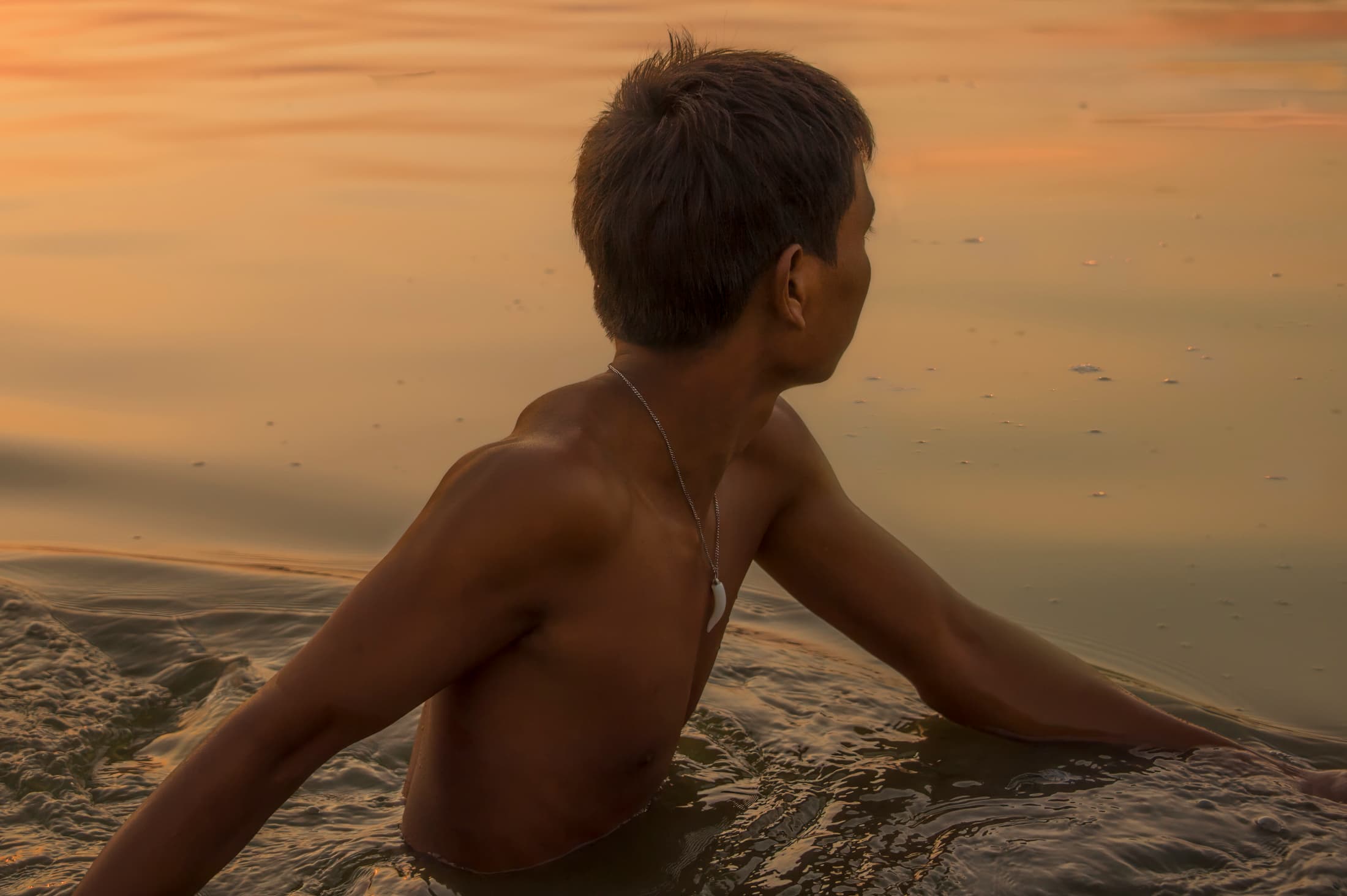 Mandalay, Myanmar (Burma). Young adult male turns away to look at lake water lit by sunset sky at twilight. Waters around him ripple as he turns and swishes his arms in the water.
