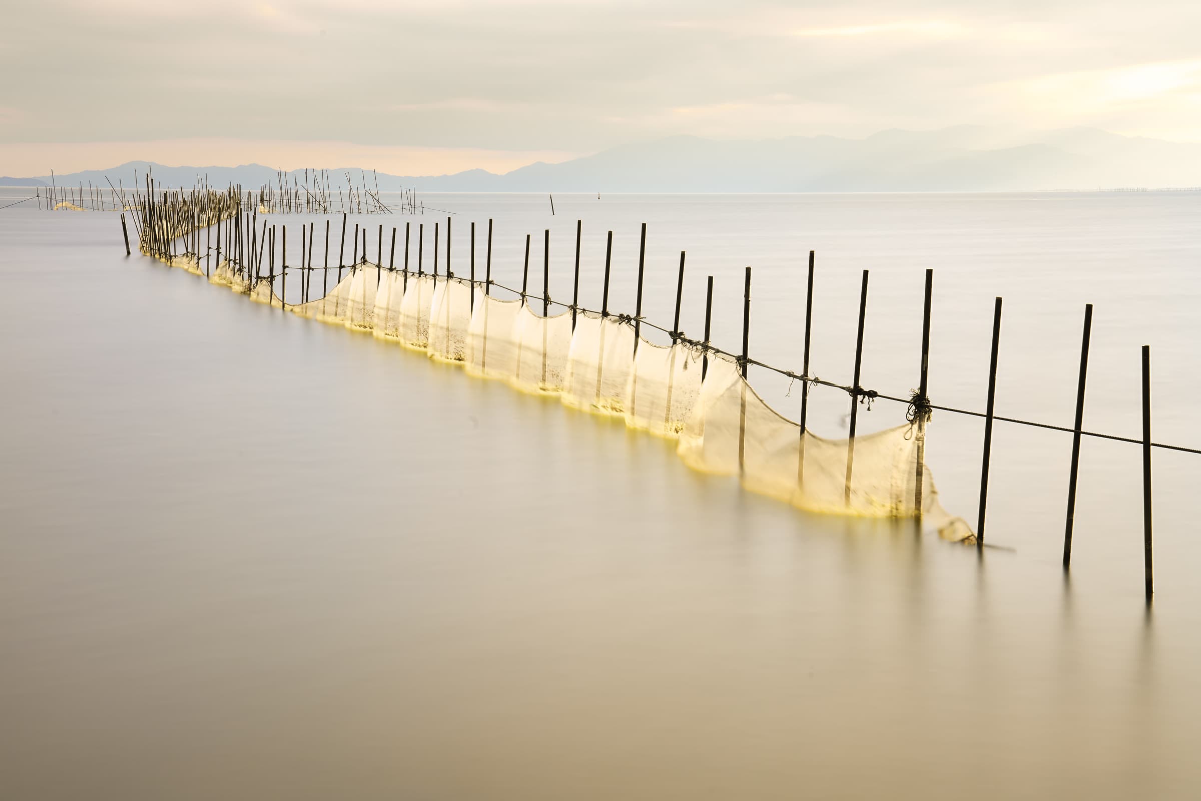 Old fishing nets and slender poles stretch across silky water in a long-exposure photograph long-exposure at Lake Biwa, glowing softly in warm yellow light.