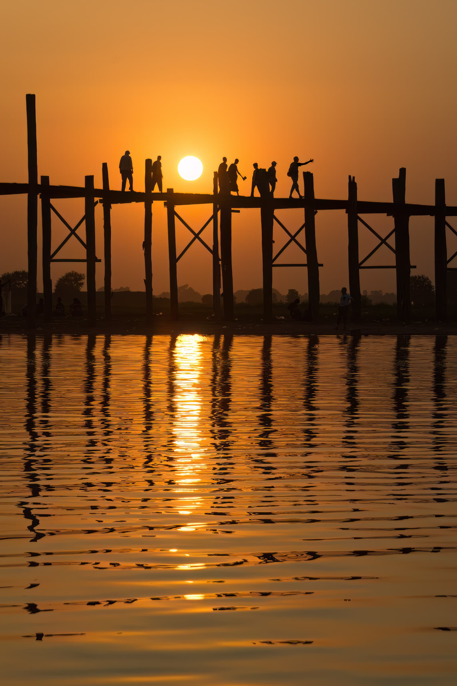 Taungthaman Lake, Mandalay,Myanmar. People walk along U Bien Bridge, a tall wooden bridge, with setting sun behind and reflections on lake water in foreground.